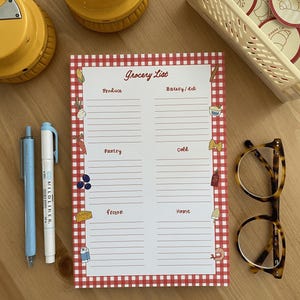 May include: A red and white gingham grocery list notepad with sections for produce, bakery/deli, pantry, cold, frozen, and home. The notepad has small illustrations of food items on each section.