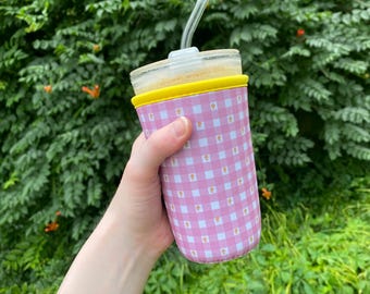 Könnte beinhalten: Ein Glasbecher mit einer gelben Silikonhülle und einer rosa Vichy-Musterhülle. Der Becher hat einen durchsichtigen Glasstrohhalm und ist mit einem hellfarbigen Getränk gefüllt. Der Hintergrund ist ein üppiger grüner Busch mit orangefarbenen Blüten.