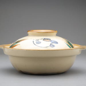 以下が含まれることがあります： ベージュ色の陶器製の日本の土鍋、donabe。 蓋には、青、緑、オレンジ色のアクセントで描かれた魚のデザインがあります。 鍋は丸い形で、2つの取っ手が付いています。