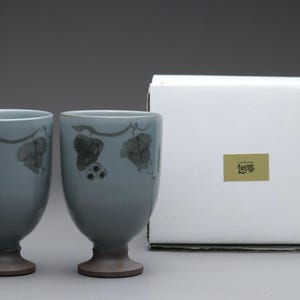 以下が含まれることがあります： ブドウの木の模様が入った、濃い茶色のベースと淡い青色のセラミックゴブレット2つ。ゴブレットは、金のラベルが付いた白い箱の隣にあります。ブドウの木の模様には、葉とブドウが含まれています。