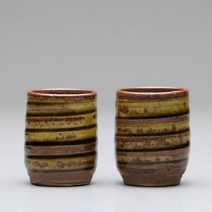 以下が含まれることがあります： 手作りのセラミックタンブラー2個。各タンブラーは、黄色、茶色、黒の水平バンドを備えた円筒形をしています。リムとベースは濃い茶色です。タンブラーはおそらく飲用です。
