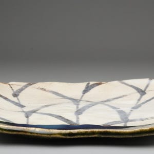 以下が含まれることがあります： クリーム色のベースと青灰色の線の模様が特徴の長方形のセラミック製サービング皿。縁は緑と茶色のトリムです。この手作りの皿は、前菜や小皿の提供に適しています。
