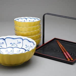 以下が含まれることがあります： 日本のスタイルの食器のコレクション。青と白の花柄の黄色いボウル、赤い縁の黒いトレイ、箸が含まれています。ボウルは積み重ねられています。