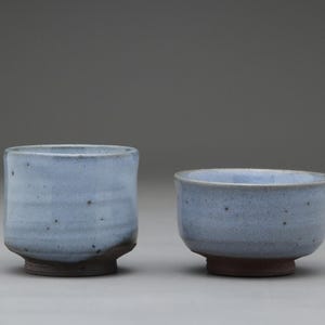 以下が含まれることがあります： 手作りの陶器のカップが2つ。カップは薄い青色で、濃い茶色の底があります。左側のカップは背が高く円筒形で、右側のカップは短く幅広です。どちらもまだらのテクスチャです。