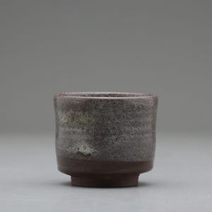 以下が含まれることがあります： 手作りの小さなセラミックカップ。表面はざらざらとしたまだら模様。カップは濃い茶色の底と、微妙な色の変化があるグレーブラウンの上部が特徴です。カップは円筒形で、縁がわずかに広がっています。