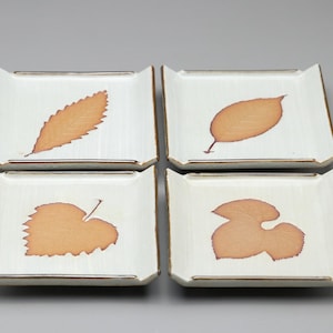 以下が含まれることがあります： 白い背景に茶色の葉の模様が描かれた、四角い陶器のプレート4枚。各プレートには異なる葉の模様があります。プレートの縁は茶色で、銀色のトリムが付いています。