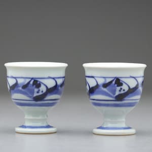 以下が含まれることがあります： 上部に青と黒の波模様が入った、小さな白い陶器のカップが2つ。カップは台座付きで、明るい灰色の表面に置かれています。
