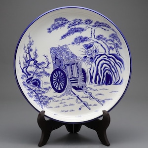 May include: A round white ceramic plate with a blue rim, featuring a detailed blue ink illustration of a traditional rickshaw centered among pine trees, flowering branches, and rock formations. The plate is displayed on a black plastic stand.