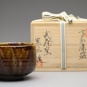 以下が含まれることがあります： 手作りの陶器の抹茶碗。外側は質感のある濃い茶色で、内側は明るく釉薬がかけられています。 抹茶碗の後ろには、日本の書道とリボンが付いた木箱があります。