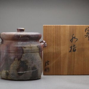 以下が含まれることがあります： 蓋と2つの持ち手が付いた円筒形の陶器製容器で、茶色と灰色のまだら模様の釉薬が施されています。容器の横には、前面に黒い日本語の書道文字が書かれた正方形の木製収納箱が置かれています。