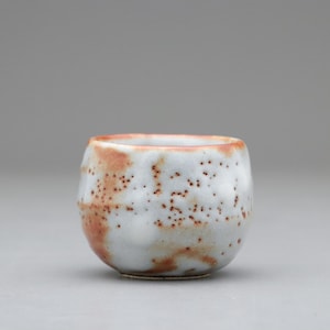 以下が含まれることがあります： 丸みを帯びた形の小さな手作りのセラミックカップ。カップは主に白で、小さな茶色の点の斑点模様があります。縁とカップの一部は赤茶色の色合いです。