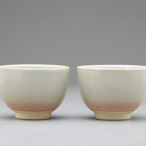 以下が含まれることがあります： クリーム色の上面とピンクがかった赤色のグラデーションが特徴の、2つのセラミックティーボウル。ボウルはシンプルで丸みを帯びた形状で、薄いベージュ色の台座の上に置かれています。背景は無地のニュートラルグレーです。