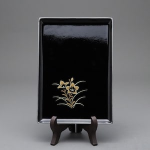 Könnte beinhalten: Ein rechteckiges, schwarzes Tablett mit silberner Umrandung und goldenem Blumendesign. Das Tablett steht auf einem dunkelbraunen Holzständer vor grauem Hintergrund. Das Blumendesign zeigt zwei Blüten und mehrere Blätter.