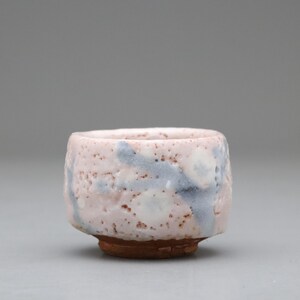 以下が含まれることがあります： 手作りの小さなセラミックボウル。表面は質感があります。ボウルは、薄いピンク色のベースに青と白のアクセントが施されています。ベースは濃い茶色です。このユニークな作品は、少量の料理を出すのに最適です。