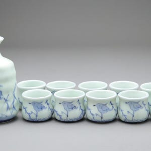 以下が含まれることがあります： ボトルと8つの小さなカップを備えた陶器の酒器セット。セットは薄い青色で、青い鳥と竹の手描きのデザインが施されています。ボトルは丸みを帯びたボディと細い首をしています。
