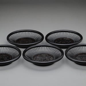 以下が含まれることがあります： 暗い茶色の、スラットデザインの丸い織りバスケットが5つ。バスケットは、中央に装飾的な模様がある浅いボウルの形をしています。バスケットは灰色の背景に並んでいます。