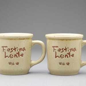 以下が含まれることがあります： ベージュ色の取っ手付きセラミックマグカップ2つ。各マグカップには、茶色の筆記体で「Festina Lente」の文字と漢字が書かれています。マグカップは斑点模様で、少し丸みを帯びた形をしています。