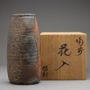 以下が含まれることがあります： まだら模様の茶色、オレンジ、青灰色の仕上げが施された、質感のある円筒形のセラミック花瓶。その隣には、日本の書道が施された木箱があります。花瓶は首が細く、ざらざらとした有機的な質感です。
