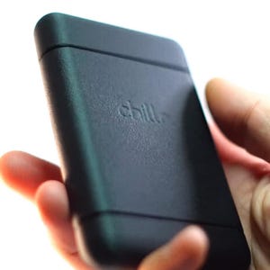 Peut inclure: Un appareil rectangulaire noir aux bords arrondis, tenu dans une main. Le mot "chill" est subtilement gravé sur la surface. L'appareil semble être fait d'un matériau mat.