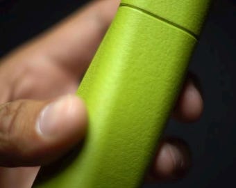 Può includere: Un astuccio rettangolare verde lime con una superficie testurizzata e una cucitura orizzontale vicino alla parte superiore, dotato di un coperchio che si separa dal corpo principale. L'oggetto ha una finitura opaca e una forma leggermente affusolata.