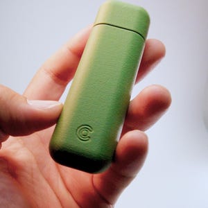 May include: A rectangular, grass-green, textured case with rounded corners and a horizontal seam near the top. The lower section features a small, embossed circular logo consisting of three concentric rings centred on the surface.