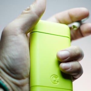 May include: A lime green rectangular container with rounded edges, held in a hand. The container has a small circular logo on the front. A small object is held in the other hand.