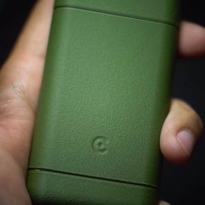Puede incluir: Un encendedor rectangular de color verde oliva con un acabado mate texturizado. El dispositivo presenta una costura horizontal cerca de la parte superior e inferior, y un pequeño logotipo circular en relieve en el centro inferior de la parte frontal.