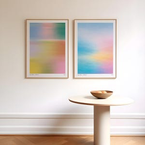 以下が含まれることがあります： ピンク、青、黄色、緑の柔らかなパステルカラーの抽象画が2枚、白い壁に額装されて飾られています。その下に、木製のフルーツボウルが置かれた、小さく丸いクリーム色のテーブルがあります。