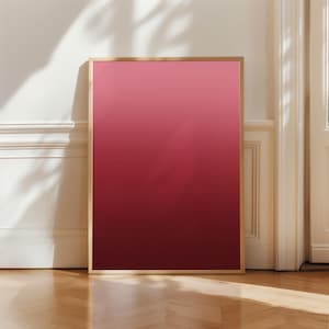 Può includere: Una stampa artistica incorniciata con un gradiente di rosa a tonalità rosso intenso. L'opera d'arte è esposta in una cornice color oro e si appoggia a una parete bianca e un pavimento in legno, creando un'estetica moderna.