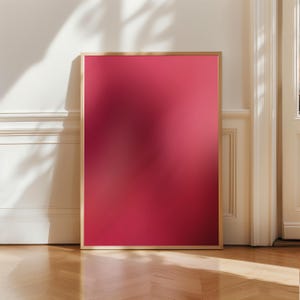 May include: A framed artwork with a vibrant, deep red gradient. The frame is a light wood color, and the artwork leans against a white wall. The floor is made of light-colored wood. The artwork is a modern, minimalist piece.