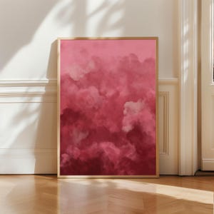 May include: A framed art print featuring a watercolor painting of pink clouds. The artwork has a gradient of colors, from light pink to deep red. The frame is a light wood color, and the print is leaning against a white wall on a wooden floor.