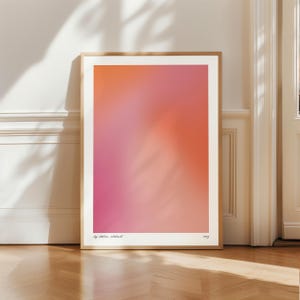 Peut inclure: Une affiche encadrée présentant un dégradé de couleurs rose et orange. L'œuvre est exposée sur un sol en bois contre un mur blanc. L'impression a un bord blanc et un fin cadre en bois. Le texte "by atelier abstract" et l'année "2023" sont imprimés en bas.