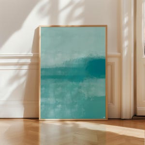 Puede incluir: Impresión artística abstracta en un marco de madera. La obra presenta una mezcla de tonos turquesa y verde azulado, con texturas de pinceladas que crean un efecto en capas. El arte enmarcado se exhibe contra una pared blanca y un suelo de madera.