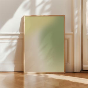Op de afbeelding: Een ingelijste kunstdruk met een kleurengradiënt, die overgaat van lichtbeige naar lichtgroen. De houten lijst omringt het kunstwerk, dat tegen een witte muur leunt. De vloer is gemaakt van houten planken.