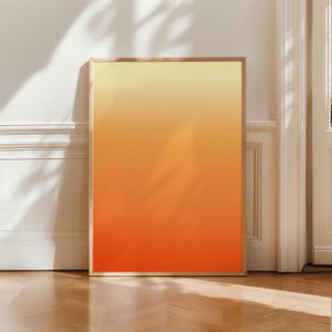 Può includere: Una stampa artistica incorniciata con una sfumatura di colore, che passa dal giallo in alto all'arancione e al rosso in basso. L'opera d'arte è esposta contro un muro bianco e un pavimento in legno.