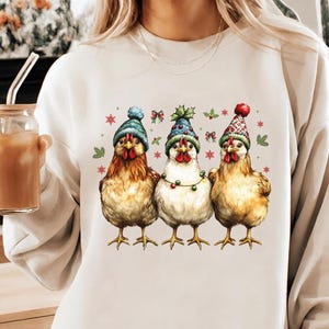 May include: A cream-colored sweatshirt featuring a graphic of three chickens wearing festive hats and a string of lights. The chickens are in shades of brown and white, with red, blue, and green accents. The sweatshirt is a casual, comfortable style.