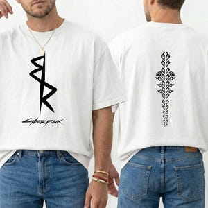 May include: White t-shirt with a black graphic design on the front and back. The front features an abstract symbol and the word "CYBERPUNK" below it. The back has a vertical floral design. The shirt is worn with blue jeans.