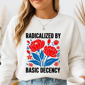 Op de afbeelding: Witte sweatshirt met de tekst "RADICALIZED BY BASIC DECENCY" boven een bloemenontwerp met rode bloemen, blauwe bladeren en rode accenten. De sweatshirt wordt door een persoon gedragen.