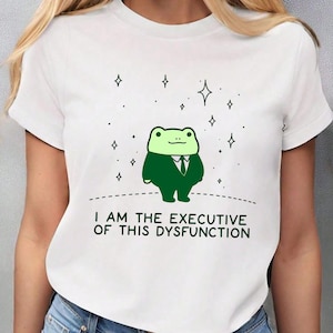 Puede incluir: Camiseta blanca con un dibujo de una rana de dibujos animados con traje, y el texto "I AM THE EXECUTIVE OF THIS DYSFUNCTION". La rana es verde con un traje y corbata negros. Pequeños gráficos de estrellas encima.