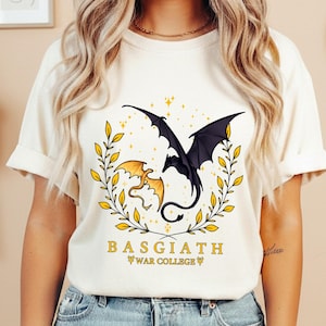 May include: Cream-colored t-shirt featuring a graphic of two dragons, one black and one gold, within a gold leaf wreath. The text "BASGIATH WAR COLLEGE" is printed below the dragons in a serif font.