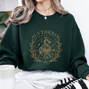 May include: Dark green sweatshirt featuring a gold-colored Slytherin Hogwarts crest. The crest includes a snake, stars, and botanical elements. The sweatshirt is a crew neck style.