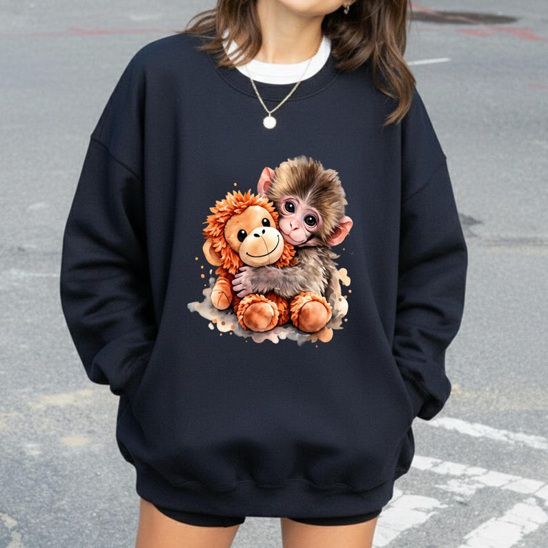 May include: Navy blue sweatshirt featuring a colorful illustration of two hugging monkeys. One monkey is brown and the other is orange. The graphic is centered on the front of the sweatshirt. The sweatshirt has a crew neck and long sleeves.