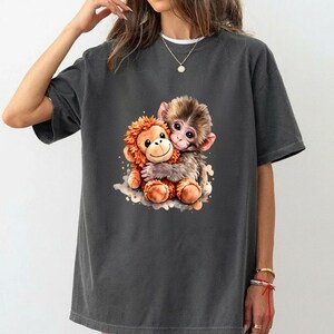 May include: Dark gray t-shirt featuring a colorful illustration of two hugging monkeys. One monkey is orange with a fluffy mane, and the other is brown with a sweet expression. The design is centered on the shirt.