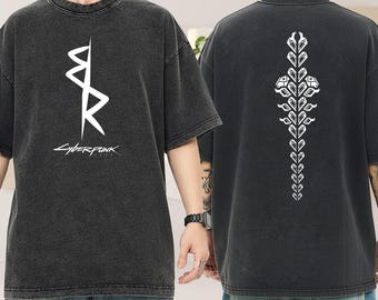 以下が含まれることがあります： 前面と背面に白いグラフィックが入った濃いグレーのTシャツ。前面には「Cyberpunk」の文字が入った様式化されたデザインがあります。背面には垂直のフローラルデザインが表示されています。シャツはヴィンテージ感のあるウォッシュ加工が施されています。