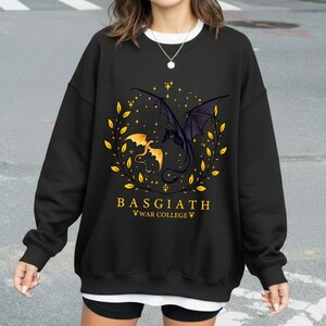 May include: Black sweatshirt featuring a dragon graphic with the text "BASGIATH WAR COLLEGE". The design includes two dragons, one gold and one black, surrounded by a wreath of yellow leaves and stars.