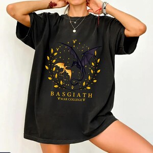 May include: Black t-shirt featuring a graphic of two dragons facing each other within a golden wreath. The text "BASGIATH WAR COLLEGE" is printed below the dragons in gold. The shirt has short sleeves.