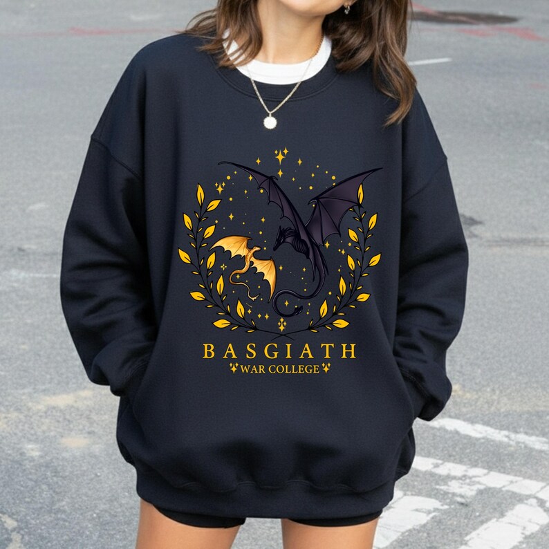 May include: Navy blue sweatshirt featuring a dragon design with a gold and black color scheme. The design includes two dragons, a laurel wreath, and the text "BASGIATH WAR COLLEGE". The sweatshirt is a comfortable, casual style.