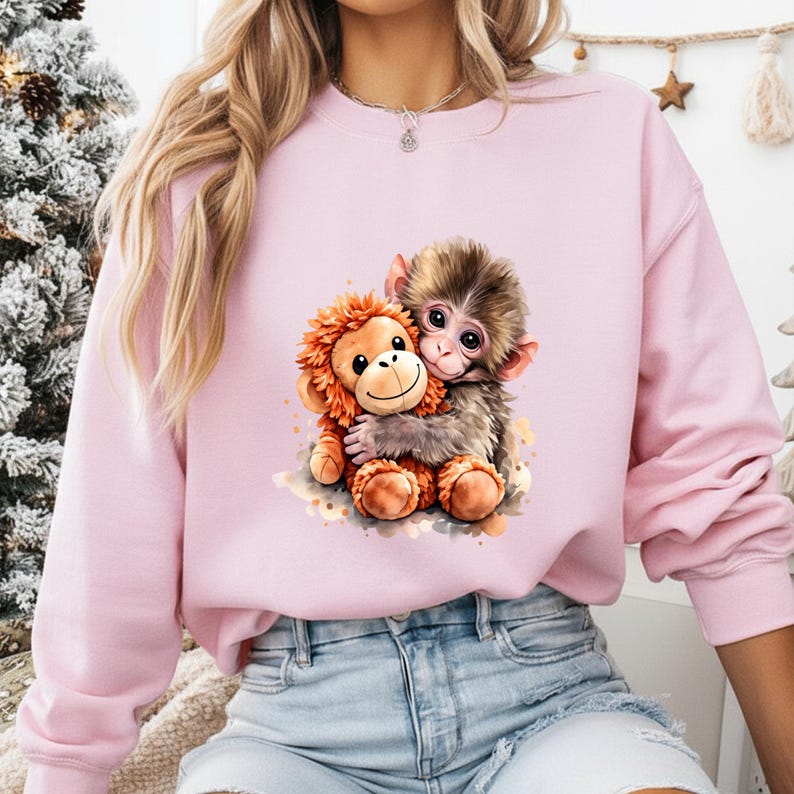 May include: A light pink sweatshirt featuring a watercolor illustration of two hugging monkeys. One monkey is brown and the other is orange. The design is centered on the front of the sweatshirt.