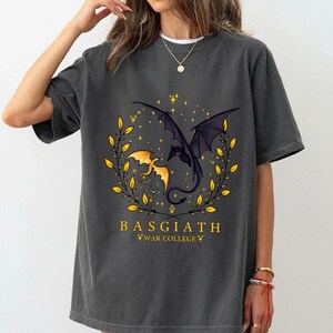 May include: Dark gray t-shirt featuring a gold and black dragon design within a laurel wreath. The text "BASGIATH WAR COLLEGE" is printed in gold below the dragons. The shirt has short sleeves.
