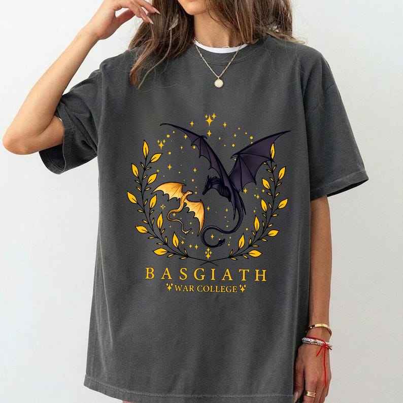 May include: Dark gray t-shirt featuring a gold and black dragon design within a laurel wreath. The text "BASGIATH WAR COLLEGE" is printed in gold below the dragons. The shirt has short sleeves.